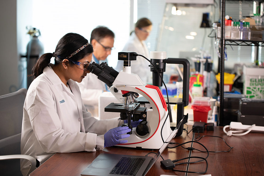 A young woman in a white lab coat looks into a microscope while two more employees in the background work on research in the Noblis applied sciences lab.