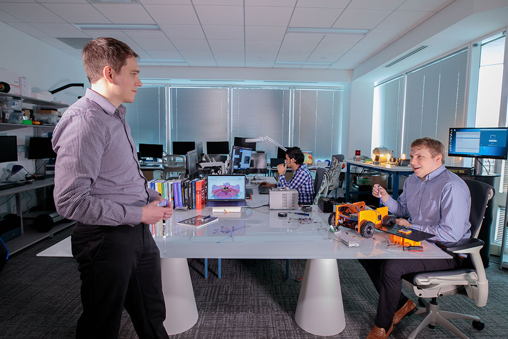 Three employees talk and work around a table with rovers and other parts for equipment research in the Noblis Autonomy and Internet of Things lab.