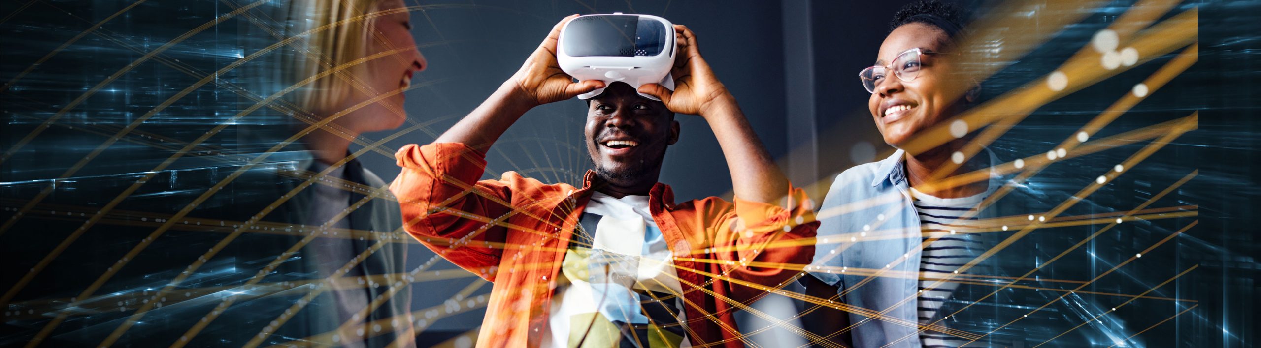 Three interns sit together in an office room. The young blonde woman smiles at a young Black man who is also smiling and holding virtual reality glasses above his face. The other young woman sits beside them also smiling.